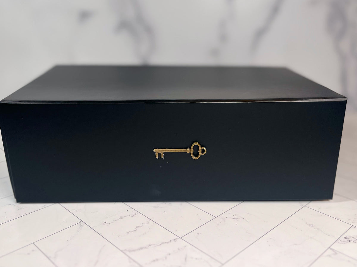 A pristine black rectangular box, featuring a gold key emblem on its front, rests atop a white marble surface streaked with gray veining, conjuring the enigmatic allure of The Murder Box Edition 100th Anniversary collector’s set.