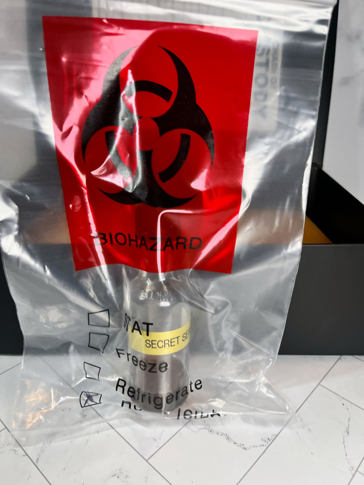 A sealed clear bag bearing a biohazard symbol holds a vial labeled “SECRET SERUM” along with storage instructions such as “Refrigerate,” displayed on white tiles—a mysterious collectible from The Murder Box Edition, ideal for enthusiasts of intriguing mysteries.