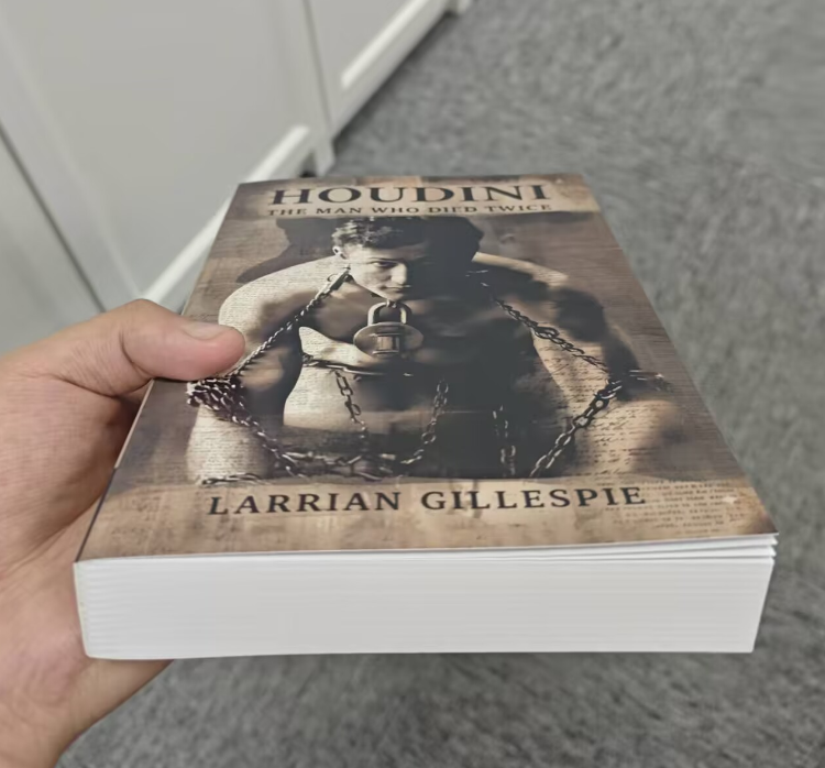 A hand holds the Trade Paperback Edition of "Houdini: The Man Who Died Twice" by Larrian Gillespie, featuring Houdini in chains on the cover—an intriguing true crime investigation into his mysterious death.