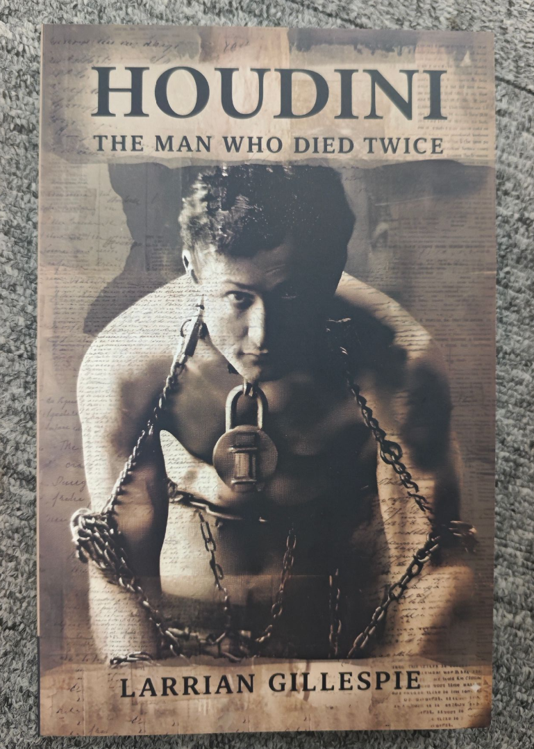 Trade Paperback Edition of "Houdini: The Man Who Died Twice" by Larrian Gillespie, with a grayscale cover showing Houdini in chains and a padlock around his neck, evoking a gripping true crime investigation into his mysterious death.