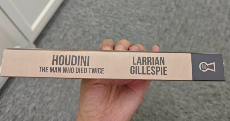 A hand holds the Trade Paperback Edition of Larrian Gillespie's "Houdini: The Man Who Died Twice," its beige spine and black text hinting at a gripping true crime investigation into Houdini's mysterious death.