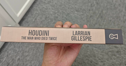 A hand holds the Trade Paperback Edition of Larrian Gillespie's "Houdini: The Man Who Died Twice," its beige spine and black text hinting at a gripping true crime investigation into Houdini's mysterious death.